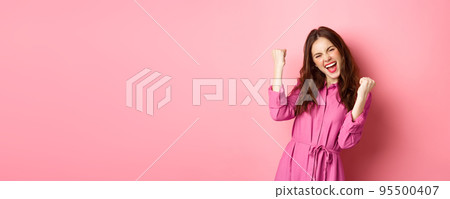 Beautiful smiling woman scream with happy and excited face, saying yes, making fist pump, winning and feeling like champion, triumphing, standing over pink background 95500407