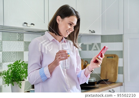 Young woman at kitchen looking in smartphone, with glass of water 95502210