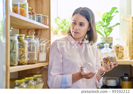 Young woman in kitchen with containers jars of food, holding jar of almonds Young woman in kitchen with containers jars of food, holding jar of almonds 95502212