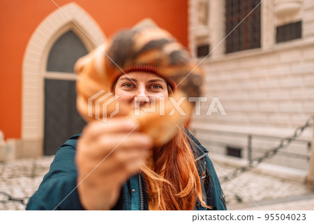 Happy turist woman in a bright hat and autumn jacket holding baked obwarzanek traditional polish cuisine snack bagel on old city Market square in Krakow Happy turist woman in a bright hat and autumn jacket holding baked obwarzanek traditional polish cuisine snack bagel on old city Market square in Krakow 95504023