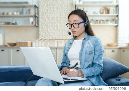 Work online. A young Asian woman is a call center operator. Sitting on the sofa at home wearing headphones with a microphone and a laptop, answering calls, consulting, writing down a request. Work online. A young Asian woman is a call center operator. Sitting on the sofa at home wearing headphones with a microphone and a laptop, answering calls, consulting, writing down a request. 95504996