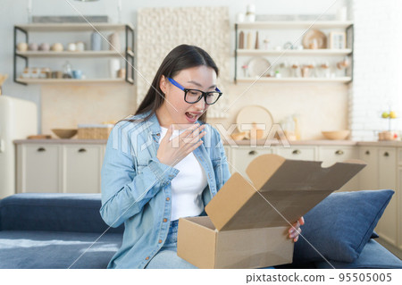 Happy and pleasantly surprised young beautiful Asian woman sitting on sofa at home and opening cardboard box, gift. I received an unexpected package in the mail. 95505005