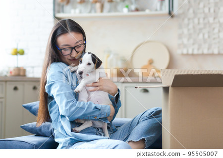 An unexpected pleasant gift. A happy and impressed young Asian woman received a small dog as a gift for her birthday. He sits at home on the sofa near a large cardboard box, rejoices, hugs the dog. An unexpected pleasant gift. A happy and impressed young Asian woman received a small dog as a gift for her birthday. He sits at home on the sofa near a large cardboard box, rejoices, hugs the dog. 95505007