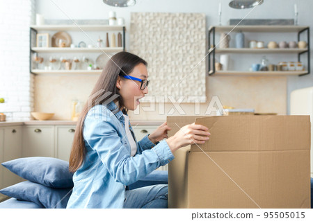 Parcel home delivery. Happy young beautiful Asian woman received parcel. Excited by surprise, she opens a huge cardboard box while sitting on the couch at home. 95505015
