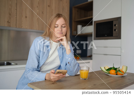 Pretty young blonde woman using smartphone and drinking orange juice while sitting at the kitchen, girl is messaging online on the phone in the morning Pretty young blonde woman using smartphone and drinking orange juice while sitting at the kitchen, girl is messaging online on the phone in the morning 95505414