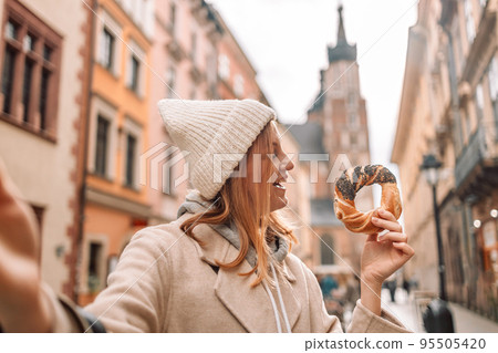 Happy young blonde woman tourist in stylish clothes holding pretzel obwarzanek on the market square in Krakow in Poland Happy young blonde woman tourist in stylish clothes holding pretzel obwarzanek on the market square in Krakow in Poland 95505420