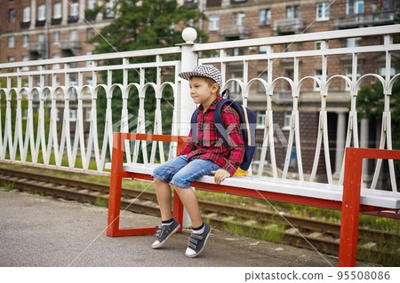 cute caucasian boy in checkered shirt, cap and jean shorts sitting on bench at rail station waiting for the train with pensive face. cute caucasian boy in checkered shirt, cap and jean shorts sitting on bench at rail station waiting for the train with pensive face. 95508086