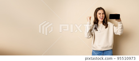Happy girl showing mobile phone horizontal screen and celebrating, chanting with joyful face, standing over beige background 95510973