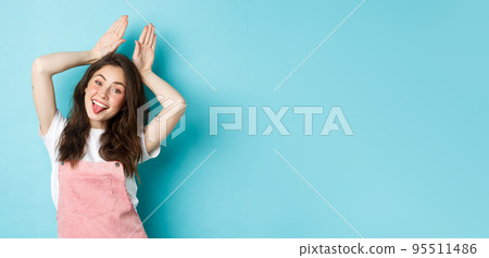 Portrait of beautiful glamour girl celebrating Easter, showing bunny rabbit ears with hands above head, smiling joyful, standing over blue background 95511486