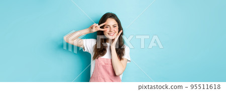 Portrait of kawaii teenage girl giggle and showing peace v-sign near eye, smiling cheerful and touching cheek, standing over blue background Portrait of kawaii teenage girl giggle and showing peace v-sign near eye, smiling cheerful and touching cheek, standing over blue background 95511618