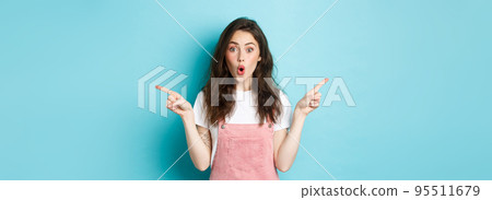 Wow check it out. Excited brunette girl with glamour summer make up, pointing fingers sideways at left and right sides, gasping amazed, standing over blue background 95511679