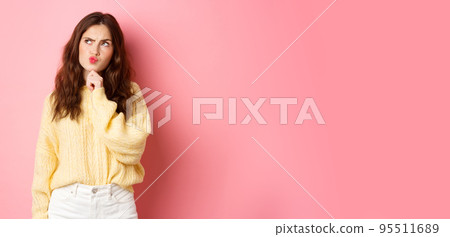 Puzzled cute girl thinking, touching chin and frowning thoughtful, looking aside at logo banner with pensive face, standing over pink background 95511689