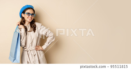 Stylish happy girl on shopping. Portrait of modern woman with shop bag, laughing and smiling satisfied, buying herself gift, standing over beige background 95511815
