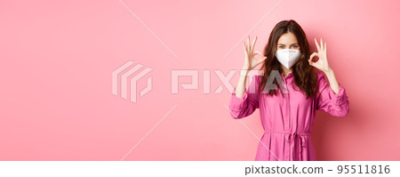 Covid-19, pandemic and lifestyle concept. Cheerful girl shows okay sign, wears medical respirator as preventive measure from corona, pink background 95511816