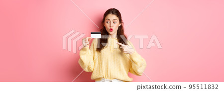 Image of glamour girl going shopping, looking excited and pointing at plastic credit card, standing against pink background 95511832