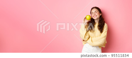 Healthcare, stomatology and people concept. Happy girl show her white perfect teeth, smile and green apple, eating healthy food and fruits, standing against pink background 95511834