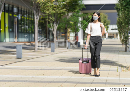 A business woman wearing a mask, carrying a backpack, and walking with a suitcase 95513357