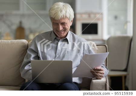 Smiling elderly man sits on sofa using laptop, holding papers Smiling elderly man sits on sofa using laptop, holding papers 95515939