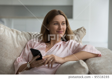 Woman looks aside sits on sofa with smartphone in hands Woman looks aside sits on sofa with smartphone in hands 95516193