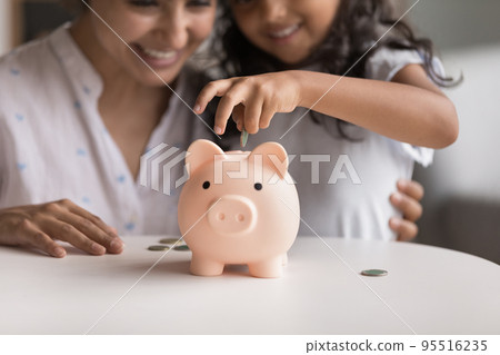 Smiling Indian mom and little daughter dropping coins into piggybank Smiling Indian mom and little daughter dropping coins into piggybank 95516235