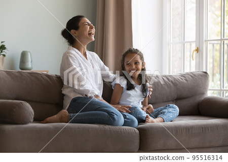 Happy Indian woman laughing seated on sofa with preschooler daughter 95516314