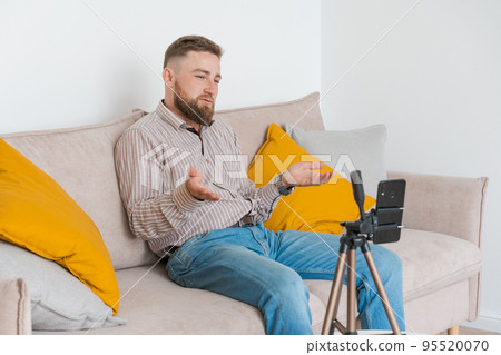 Smiling bearded guy filming video using smartphone on tripod online while 95520070