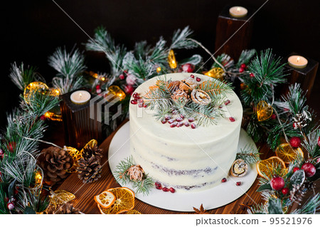 Christmas cake made of chocolate base with cream, decorated with spruce branches, walnuts and pomegranate seeds against the backdrop of Christmas decor Christmas cake made of chocolate base with cream, decorated with spruce branches, walnuts and pomegranate seeds against the backdrop of Christmas decor 95521976