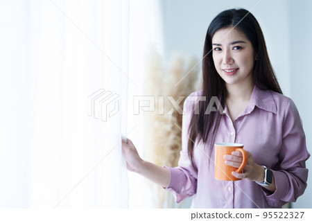 A portrait of a young Asian woman in pajamas smiling happily by a window with curtains in the morning sun while drinking coffee. A portrait of a young Asian woman in pajamas smiling happily by a window with curtains in the morning sun while drinking coffee. 95522327