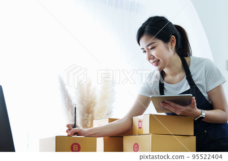 Starting small business entrepreneur of independent Asian woman smiling using tablet computer with cheerful success of online marketing package box items and SME delivery concept. 95522404