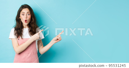Impressed brunette girl pointing fingers right at logo banner, staring with excitement and surprised face, telling big news, showing promo offer, blue background Impressed brunette girl pointing fingers right at logo banner, staring with excitement and surprised face, telling big news, showing promo offer, blue background 95531245