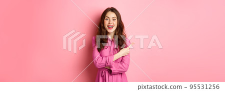 Stylish young modern woman look amazed, pointing finger left aside at copyspace, showing promo text, standing happy against pink background Stylish young modern woman look amazed, pointing finger left aside at copyspace, showing promo text, standing happy against pink background 95531256