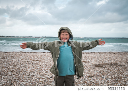 Boy is playing with sand and wind at the beach. School kid near water at autumn winter sea. Child having fun with raised hands and running. Waves at the shore. Spring Holiday vacation concept. 95534353
