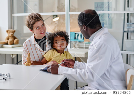 Young woman visiting pediatrician with her little son, they sitting at table and talking to doctor Young woman visiting pediatrician with her little son, they sitting at table and talking to doctor 95534865