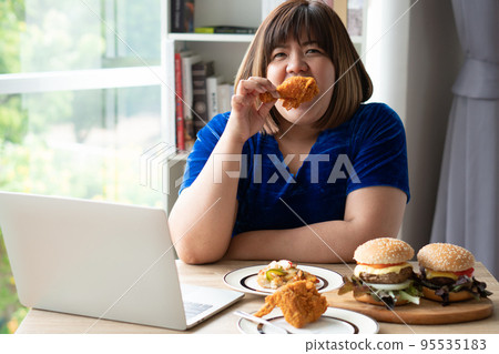 Hungry overweight woman holding Fried Chicken, hamburger on a wooden plate and Pizza on table, During work from home, gain weight problem. Concept of binge eating disorder (BED). 95535183