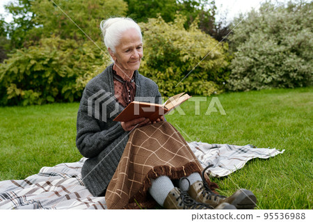 Senior woman in casualwear sitting on checkered woolen plaid and reading 95536988