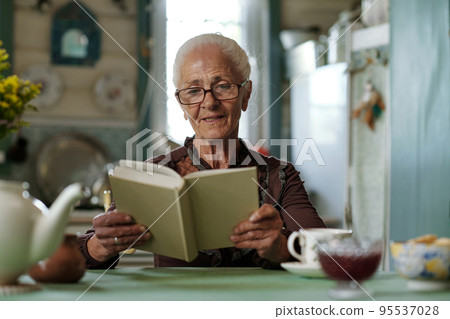 Senior woman in eyeglasses reading captivating story or novel after breakfast Senior woman in eyeglasses reading captivating story or novel after breakfast 95537028