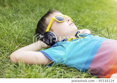 Boy in sunglasses lying on the grass and listening to music in headphones Boy in sunglasses lying on the grass and listening to music in headphones 95538124