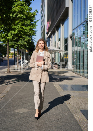 Vertical shot of businesswoman walking on street with digital tablet, going to work, wearing beige suit and high heels 95539884