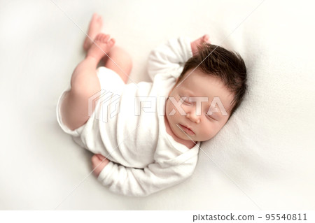 Top view of a newborn baby girl lying in a white bodysuit on a white bed. Beautiful portrait of a little sleeping girl with closed eyes 7 days, one week. Photography in macro studio. Top view of a newborn baby girl lying in a white bodysuit on a white bed. Beautiful portrait of a little sleeping girl with closed eyes 7 days, one week. Photography in macro studio. 95540811