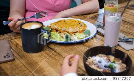Close-up of child girl with her mother having vegan breakfast at cafe or restaurant, slow motion. Woman eating vegan chickpea omelet with mushrooms while her daughter eating blueberry smoothie bowl Close-up of child girl with her mother having vegan breakfast at cafe or restaurant, slow motion. Woman eating vegan chickpea omelet with mushrooms while her daughter eating blueberry smoothie bowl 95547267