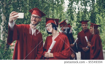 Cheerful couple of graduating students is taking selfie using smartphone, young attractive man and woman in glasses are holding diplomas, looking at smart phone camera and smiling. Cheerful couple of graduating students is taking selfie using smartphone, young attractive man and woman in glasses are holding diplomas, looking at smart phone camera and smiling. 95549394