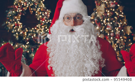 Portrait of man in traditional Santa Claus costume and glasses looking at camera near Christmas tree in dark illuminated and decorated room. Holidays and people concept. 95549408