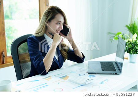 Portrait of a young Asian woman showing a smiling face as she uses her phone, computer and financial documents on her desk in the early morning hours. Portrait of a young Asian woman showing a smiling face as she uses her phone, computer and financial documents on her desk in the early morning hours. 95551227