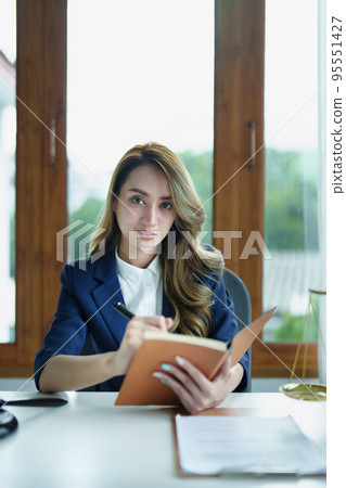 Portrait of a young Asian lawyer studying a lawsuit for a client using notebooks and paperwork on a desk. Portrait of a young Asian lawyer studying a lawsuit for a client using notebooks and paperwork on a desk. 95551427