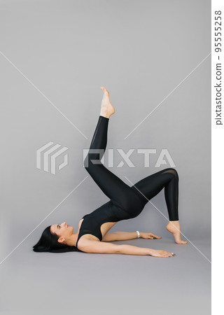 Young woman fitness trainer or yoga performs an exercise lying on her shoulder blades and lifting one leg at the top on a gray background Young woman fitness trainer or yoga performs an exercise lying on her shoulder blades and lifting one leg at the top on a gray background 95555258