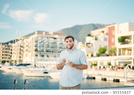 Young man with ice cream in his hands stands on the embankment with moored yachts Young man with ice cream in his hands stands on the embankment with moored yachts 95555837
