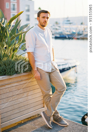 Young man stands on a pier leaning against a wooden tub with green plants Young man stands on a pier leaning against a wooden tub with green plants 95555965