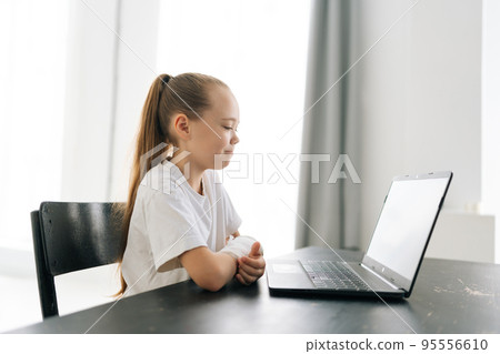 Side view of cute blonde little girl with broken hand wrapped in white plaster bandage sitting at table with laptop computer looking to screen in light room. Concept of child insurance and healthcare. Side view of cute blonde little girl with broken hand wrapped in white plaster bandage sitting at table with laptop computer looking to screen in light room. Concept of child insurance and healthcare. 95556610