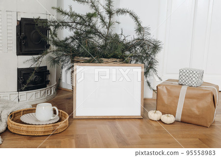 Festive Christmas home decor. Landscape wooden picture frame mockup on parquette floor. Pine, larch tree branches in basket, gift boxes. Cup of coffee. Old white tiled stove background.Empty template Festive Christmas home decor. Landscape wooden picture frame mockup on parquette floor. Pine, larch tree branches in basket, gift boxes. Cup of coffee. Old white tiled stove background.Empty template 95558983