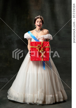 Christmas present. Young queen. Portrait of adorable girl in image of medieval royal person in renaissance style dress isolated on dark background. Comparison of eras, beauty, history, art Christmas present. Young queen. Portrait of adorable girl in image of medieval royal person in renaissance style dress isolated on dark background. Comparison of eras, beauty, history, art 95559384
